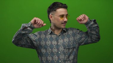 Genç İspanyol adam yeşil bir arka planda neşeli bir şekilde dans ediyor. Parlak bir modern görünüş için desenli gömlek ve eşsiz gözlük takıyor.