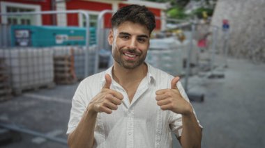 Beyaz tişörtlü adam güneş ışığı altındaki inşaat alanında duruyor. Parlak bir şekilde gülümsüyor ve bina girişinde başparmak kaldırıyor..