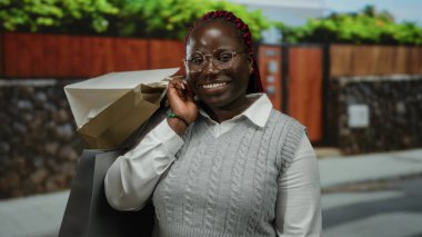 Şehir merkezindeki açık hava ortamında gözlük ve süveter takan alışveriş torbalarıyla gülen bir kadın..