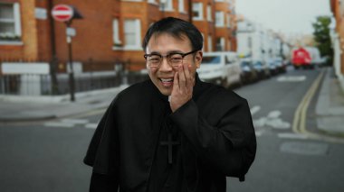 Siyah bornoz ve çapraz kolyeli gözlük takan rahip şehir ortamında neşeli bir ifade sergiliyor. Dini huzur ve neşeyi dışarıda yakalıyor.