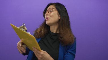 Orta yaşlı, gözlüklü bir kadın sarı pano tutuyor ve stüdyoda mor duvara uzanmış el hareketleriyle sevinç ifadesi iletişimi kuruyor..