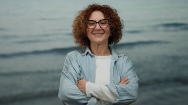 Orta yaşlı, esmer, kıvırcık saçlı ve gözlüklü İspanyol kadın kollarını çapraz bir zemine dayamış gülümsüyor, kendine güveni ve sıcaklığıyla..