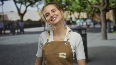 Yeşil ağaçlarla çevrili, kahverengi önlüklü kadın gündüz vakti arka planda bulanık yayalar görüyor..