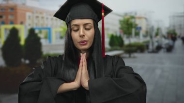 Latin kökenli genç bir kadın, mezuniyet elbisesi içinde, açık havada huzurlu bir ifadeyle akademi ve sükunetin dengeli bir karışımını sergiliyor.