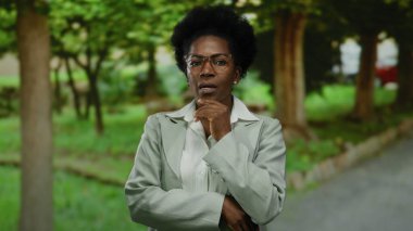 Parkta kendinden emin bir şekilde duran, etrafı ağaçlar ve çimlerle çevrili gözlükler takan kadın, açık havada sakin bir anı yakalar..