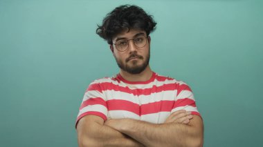 Mavi arka planda kollarını kavuşturan gözlüklü ve sakallı İspanyol genç adam çizgili gömlek giyiyor sakin bir ifade sergiliyor..