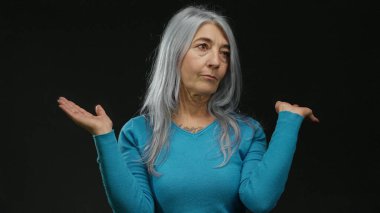 Gri saçlı ve mavi kazaklı bir kadın iki elini kaldırarak derin düşüncelere dalmış ve kafa karışıklığı olduğunu öne sürüyor..