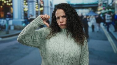 Kıvırcık saçlı, süveter giyen bir kadın, kalabalık şehir caddesinde kapalı şehir arka planında tasvip etmediğini belirterek baş parmak hareketi yapıyor..