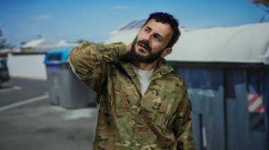 Askeri üniformalı İspanyol bir adam dışarıda çöp konteynırının yanında duruyor. Rahatsızlık içinde boynu ovalıyor, şehir ortamında stresi ya da yorgunluğu gösteriyor..