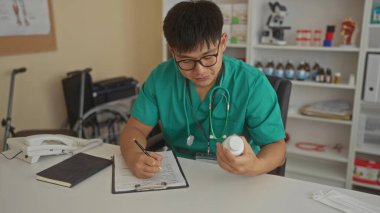 Yeşil üniformalı genç bir doktor klinikte çalışıyor, şişeyi inceliyor, not alıyor, sağlık ekipmanları ve telefon ile çevrili..