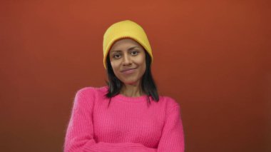 Pembe süveter ve sarı bere giyen bir kadın canlı kırmızı bir duvara karşı, gençlik cazibesi ve İspanyol havası ile modayı harmanlıyor.