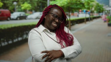 Saçları örgülü ve gözlüklü gülümseyen Afro-Amerikalı kadın şehir yeşillikleriyle çevrili ve güneşli bir günde arabaların yanından geçen şehir caddesinde kolları çapraz bir şekilde duruyor..