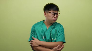 Önlüklü genç adam, gözlüklü ve steteskoplu izole edilmiş sarı arka plana karşı üşümüş hissediyor. Elleri sımsıkı bağlı, soğuk bir ortamda rahatsızlık gösteriyor.