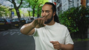 Adam, beyaz tişört giyen, çıplak elle görünen ve sakallı bir şehir caddesini işaret ediyor; özgüven odaklı..