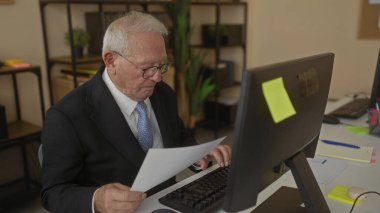 Takım elbiseli kıdemli adam, bilgisayar, notlar ve bitkilerle çevrili ofis ortamında profesyonel bir iş ortamını yansıtan belgeler okuyor..