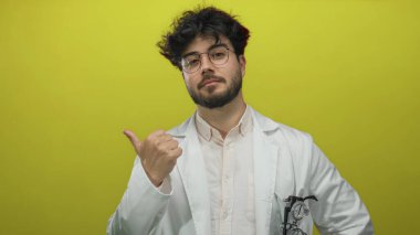 Beyaz önlüklü, sakallı ve gözlüklü genç İspanyol adam, sağlıklı bir ortamda onay ve pozitiflik öneren canlı sarı arka plana karşı emin adımlarla işaret ediyor..