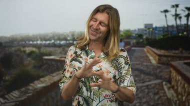 Çiçekli gömlekli, uzun sarı saçlı, kolları çaprazlanmış, parlak güneş ışığı altında yeşil bir parkta gülümseyen bir adam..