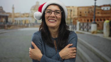 Gözlüklü ve Noel Baba şapkalı bir kadın Roma harabelerinde dururken kollarını kavuşturarak gülümsüyor..