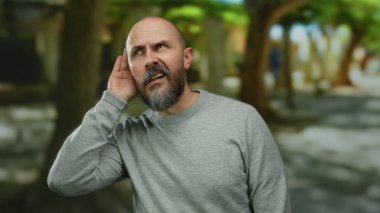 Sakallı ve kel kafalı bir adam, gri kazaklı, şehir caddesinde dikkatle dinliyor, bulanık bir şehir arka planına karşı tuhaf bir ifade sergiliyor..