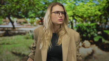 Yeşil bir parkta tutkulu bir şekilde konuşan, gözlüklü ve bej ceketli, gün boyunca yemyeşil, hareketli jestler yapan bir kadın..