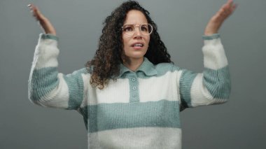 Kıvırcık saçlı ve gözlüklü bir kadın elleriyle kulaklarını kapatıyor, gri bir arka plana karşı stres ifade ediyor, çizgili bir kazak giyiyor endişeli bir ifadeyle, kapalı alanda izole edilmiş..