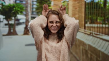 Kentsel bir sokak ortamında komik suratlar yapan, gözlüklü ve kıvırcık saçlı bir kadın sonbahar modasına uygun bir şehir ortamında neşeli bir ifade sergiliyor..