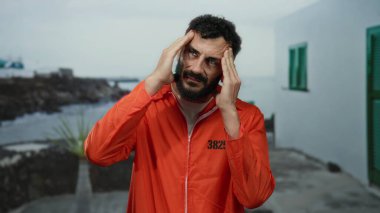 Turuncu tulumlu, sakallı İspanyol bir adam, arka plandaki kayalık bir plaja çarpan okyanus dalgaları gibi kafasında hapis ve özgürlük düşünceleri uyandırarak denizin kenarında duruyor..