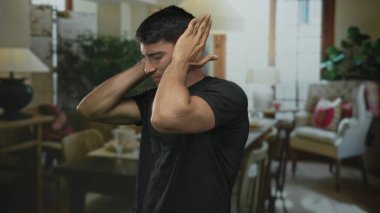 Bir bina içinde, rahat oturma odası sandalyeleri ve lamba ortasında adam iki eliyle kulakları kapatır; sıkıntı.