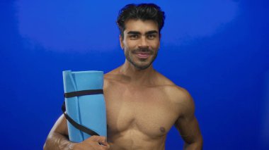 Elinde yoga minderi tutan adam parlak mavi stüdyoda çıplak göğsüne yaslanmış, kendine güveni tam sağlıklı..