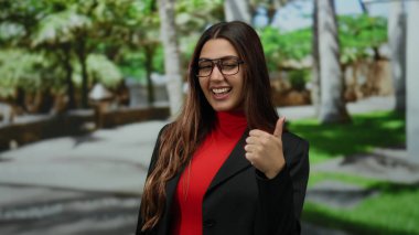 Güneşli bir parkta gülümseyen İspanyol kadın gözlüklü ve kırmızı gömlekli neşeli ve pozitif bir atmosferde ağaçlarla çevrili..