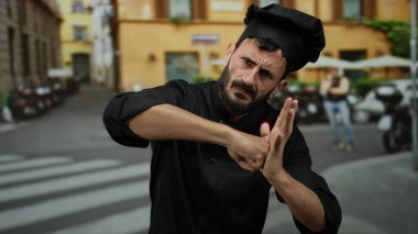 İspanyol sakallı, şef gibi giyinmiş, sokak ortamında boks pozu veren, profesyonel kıyafetleri oyuncu ifadelerle harmanlayan bir adam..