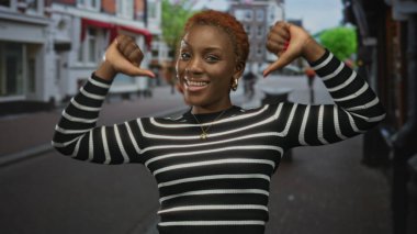 Afrika kökenli Amerikalı bir kadın kameraya gülümserken şehir caddesinde başparmağıyla göğsünü işaret ediyor..