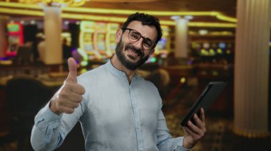 Sakallı İspanyol bir adam elinde tabletle güvenle gülümsüyor ve arka planda oyun makineleri olan canlı kumarhane ortamında başparmak kaldırıyor..