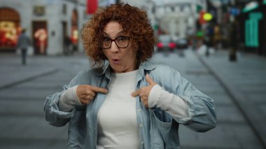 İspanyol orta yaşlı, kıvırcık saçlı ve gözlüklü bir kadın şehir caddesinde şaşırmış, günlük kıyafetler giymiş, kendini işaret ediyor..