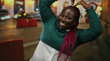 Kollarıyla gülümseyen ve kafasında kalp şekli oluşturan bir kadın. Canlı bir şehir sokak ortamında..