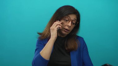 Orta yaşlı, kahverengi çerçeveli gözlüklü, mavi ceketli ve siyah bluzlu İspanyol kadın stüdyoda akıllı telefonu kulağına tutuyor..