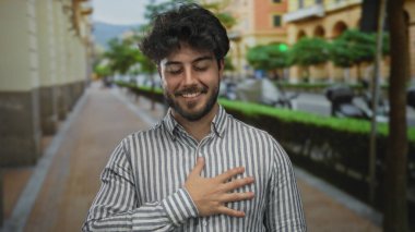 Sokakta sakallı genç bir adam, çizgili bir gömlek giyiyor, kalbinin üzerinde bir el ile gülümsüyor, bulanık yeşillik ve açık bir gökyüzünün altındaki binalarla çevrili..