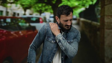 Ağaçları ve arabaları olan, kot ceketli ve sakallı, üzgün bir ifadeyle omzunu tutarak rahatsızlığını gösteren İspanyol bir adam..