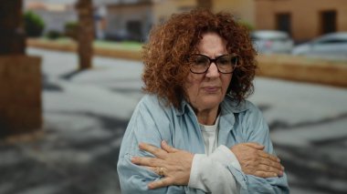 Şehir sokaklarında soğuk havada titreyen, kıvırcık saçlı ve gözlüklü mavi gömlekli bir kadın rahatsız görünüyor..