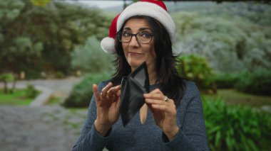 Noel Baba şapkası takan ve gözlüklü orta yaşlı bir kadın canlı yeşil bir parkta boş cüzdan tutuyor..
