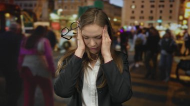 Gece vakti kalabalık bir aydınlık şehir sokağında gözlüğü yanında sallanırken, bir kadın şakaklarını iki eliyle bastırıyor..