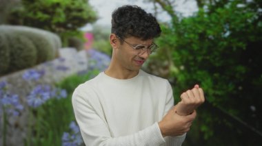 Çiçeklerin açtığı ve yeşil bir park yolunda çitleri budadığı sırada İspanyol genç adam bileğini tutuyor..