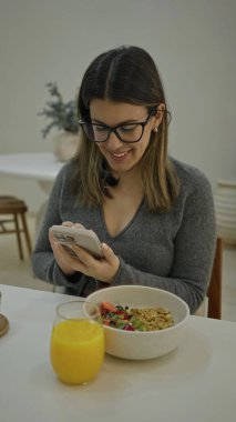 Lüks restoranda akıllı telefon kullanan bir kadın modern dekor, bir bardak meyve suyu, bir kase mısır gevreği ve şık bir ortam, sıradan bir yemek anı yakalıyor..