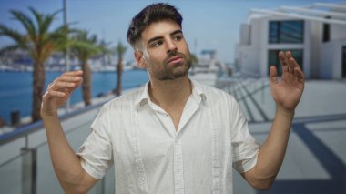 Adam sokağın terasında kollarını omuz silkerek beton bina ve deniz kenarındaki palmiye ağaçlarının yanında duruyor; kayıtsızlık.