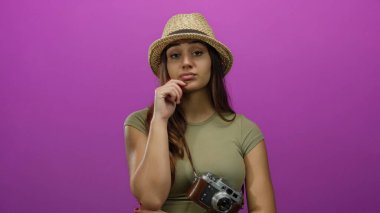 Hasır şapkalı bir kadın pembe arka planda kamera tutuyor, düşünceli bir turist havasına bürünüyor, klasik macera havası için asılı duran bir kamerayla..