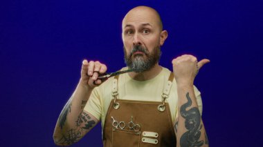 Sakallı bir erkek berber. Mavi arka planda makas tutuyor. Dövmeli, sarı tişörtlü ve deri önlüklü..