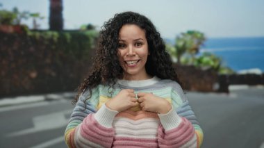 Sahilde güneşli bir günün tadını çıkaran bir kadın. Arka planda deniz var. Renkli bir kazak giyiyor..