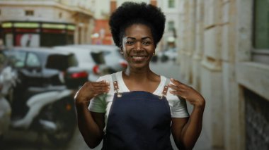 Şehir caddesinde gülen önlük giymiş bir kadın. Etrafı bulanık araba ve binalarla çevrili. Gündüz vakti şehir merkezinde..