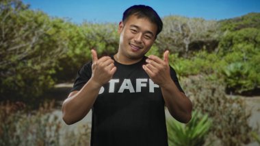 Personel tişörtlü adam yemyeşil orman yolu ve gün ışığında çalılar arasında açık elleri ile gülümsüyor ve jestler; davet bağlantısı sıcak karşılama.