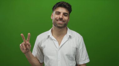 Beyaz gömlekli genç İspanyol adam stüdyoda yeşil duvarın önünde barış işareti göstererek elini tutuyor..
